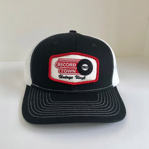 Trucker style cap with black fabric front and white mesh back, featuring a distinctive Record Town logo patch.
